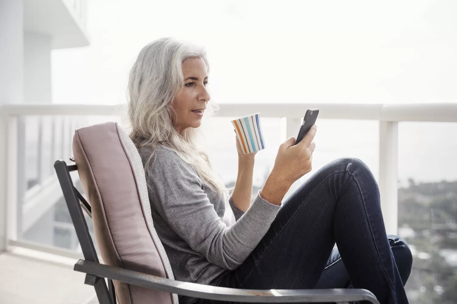 A senior woman drinks coffee on her porch, looking at her phone