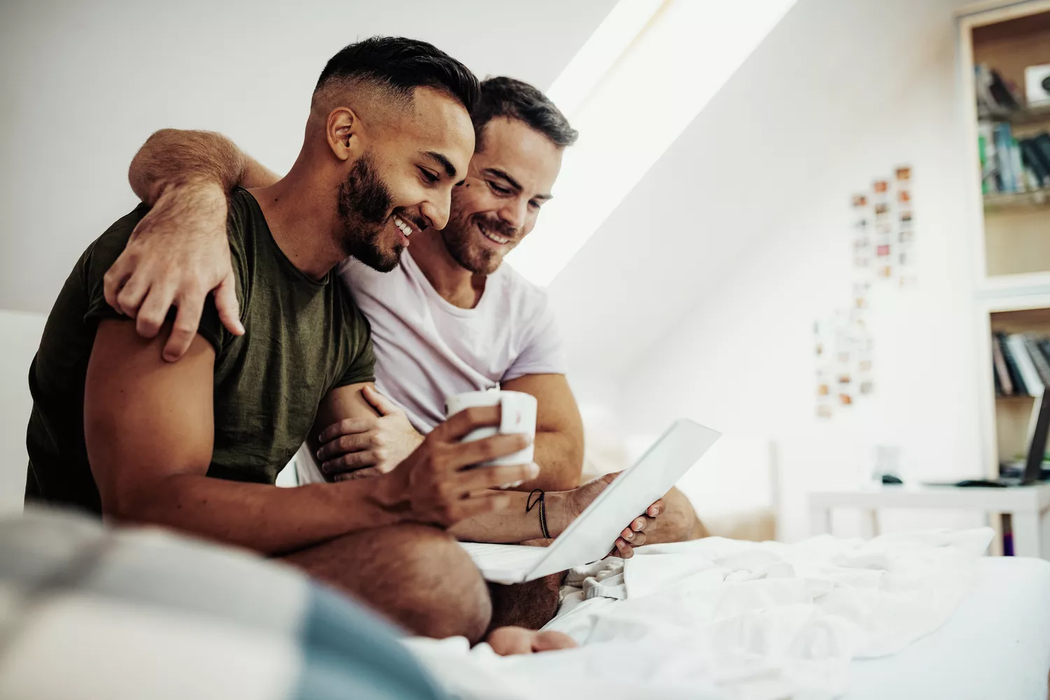 A couple looks at a laptop while sitting in bed, smiling, one with an arm around the other's shoulders